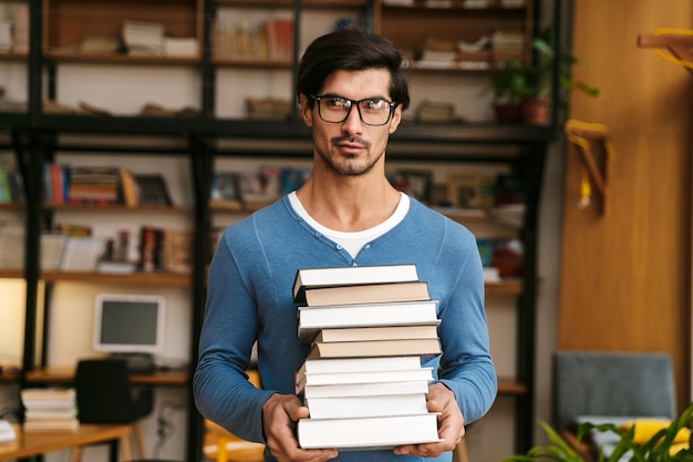 Hübscher junger Mann, der eine Brille trägt, die an der Bibliothek steht und Stapel Bücher hält