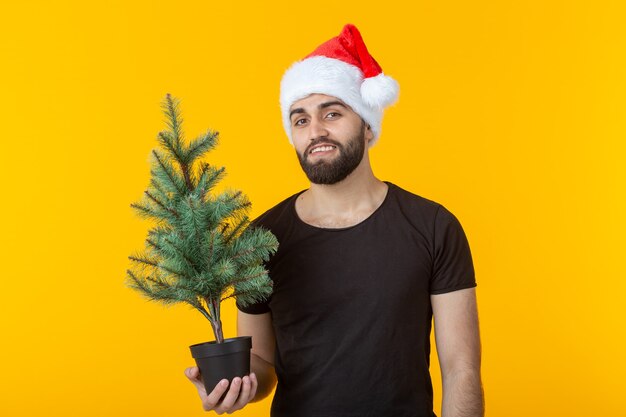 Hübscher junger Mann, der einen Weihnachtsbaum in seinen Händen hält, der im Weihnachtshut aufwirft