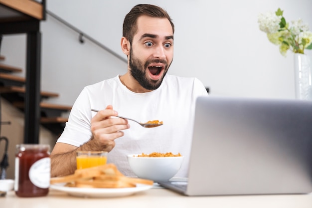 Hübscher junger Mann, der Frühstück beim Sitzen an der Küche, usinh Laptop-Computer hat