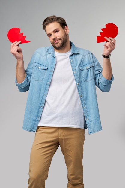 hübscher junger Mann, der gebrochenes rotes Valentinstagherz des Papiers hält, das über grau steht