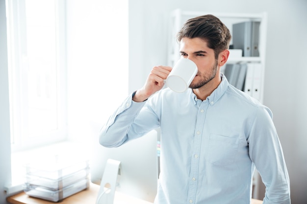 Hübscher junger Mann, der im Büro steht und Ciffee trinkt