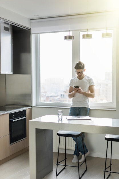 Hübscher junger Mann, der mit seinem Smartphone in der Küche eine SMS sendet