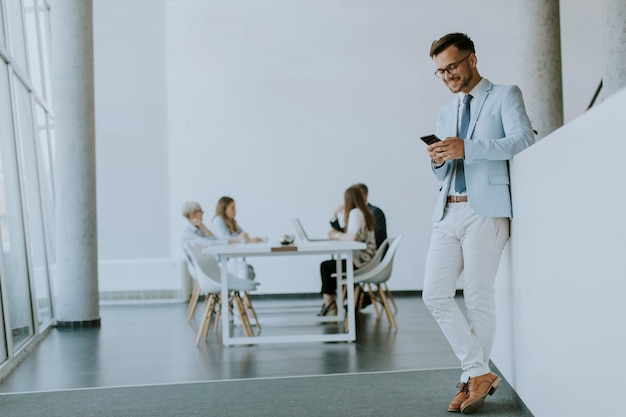 Hübscher junger Mann, der sein Handy im Büro benutzt, während seine Kollegen im Hintergrund arbeiten