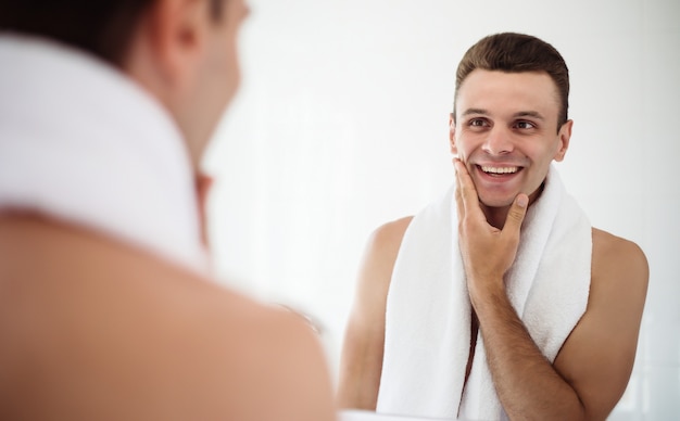 Hübscher junger Mann, der seinen Bart im Badezimmer rasiert. Porträt eines stilvollen nackten bärtigen Mannes, der sein Gesicht im Hausspiegel untersucht.