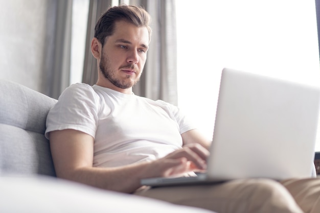 Hübscher junger Mann, der seinen Laptop mit Lächeln benutzt, während er zu Hause auf der Couch sitzt.