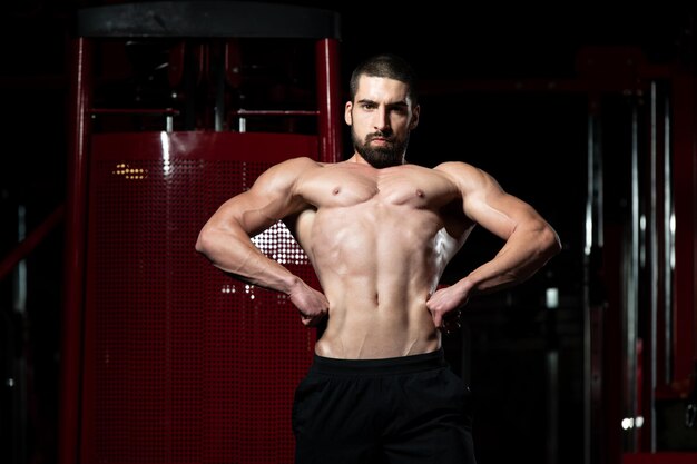 Hübscher junger Mann, der stark in der Turnhalle steht und Muskeln anspannt - muskulöser athletischer Bodybuilder-Eignungsmodell, der nach Übungen aufwirft