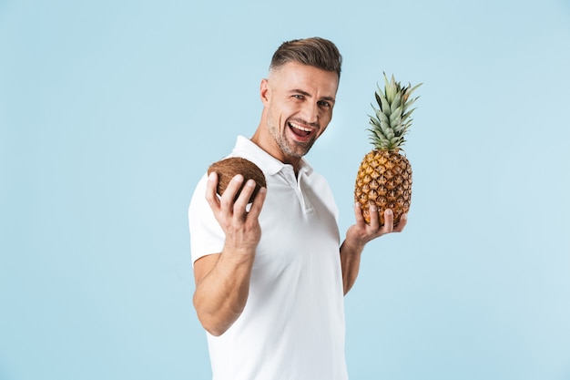 Hübscher junger Mann, der weißes T-Shirt trägt, das über Blau steht und Ananas und Kokosnuss hält