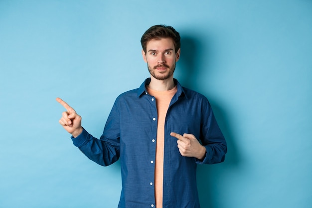 Hübscher junger Mann im Hemd, Finger links auf Promo zeigend, Logo zeigend, stehend auf blauem Hintergrund.