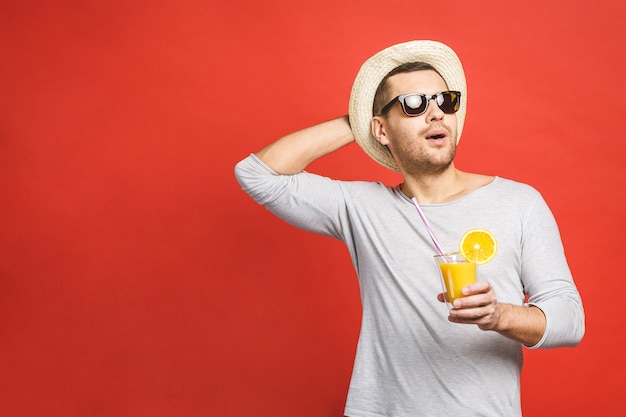 Hübscher junger Mann im Hut und in der Sonnenbrille, die frischen Orangensaft über rotem Hintergrund trinken