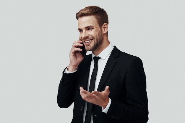 Hübscher junger Mann in Abendkleidung, der am Telefon spricht und lächelt, während er vor grauem Hintergrund steht