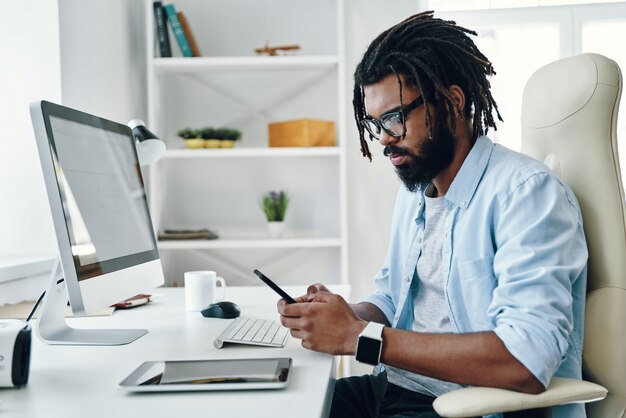 Hübscher junger Mann in Brillen mit Smartphone während der Arbeit im Büro