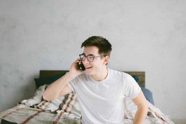 Hübscher junger Mann in der Brille, die einen Anruf macht, während auf Bett im Schlafzimmer sitzend