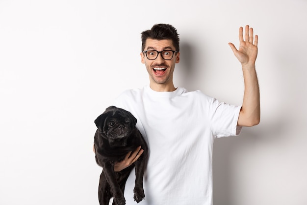 Hübscher junger Mann in der Brille, die seinen schwarzen Mops hält und Hand winkt, Mann, der Hallo sagt, während er Hund mit einem Arm trägt und über Weiß steht.