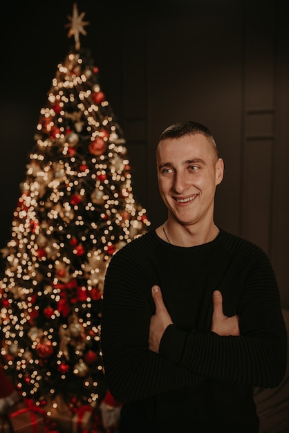 Foto hübscher junger mann in der schwarzen kleidung nahe einem weihnachtsbaum in girlanden