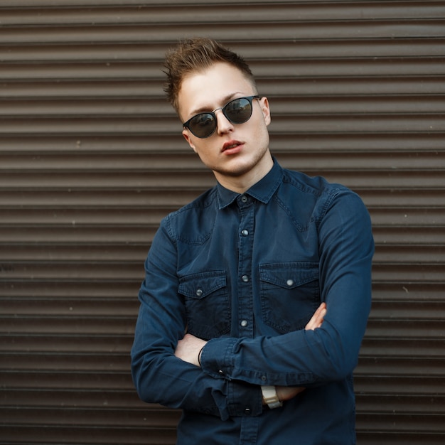 Hübscher junger Mann in einem blauen Hemd und in der Sonnenbrille nahe einer Metallwand