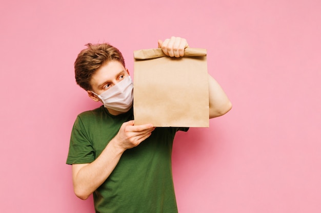 Hübscher junger Mann in einer medizinischen Schutzmaske steht auf einem Rosa mit einer Packung Lebensmittel von der Lieferung. Lieferung unter Quarantäne.