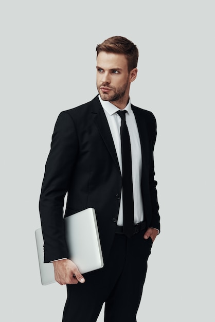 Hübscher junger Mann in formeller Kleidung, der Laptop trägt und wegschaut, während er vor grauem Hintergrund steht