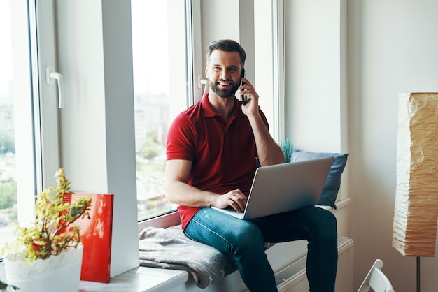 Hübscher junger Mann in Freizeitkleidung mit Laptop und Telefonieren beim Sitzen auf der Fensterbank sitting