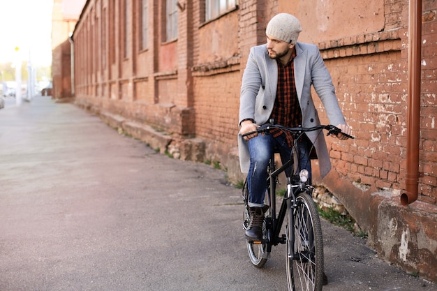 Hübscher junger Mann in grauem Mantel und Hut, der eine Fahrradstraße in der Stadt fährt.