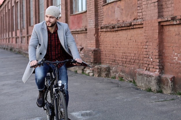 Hübscher junger Mann in grauem Mantel und Hut, der eine Fahrradstraße in der Stadt fährt.