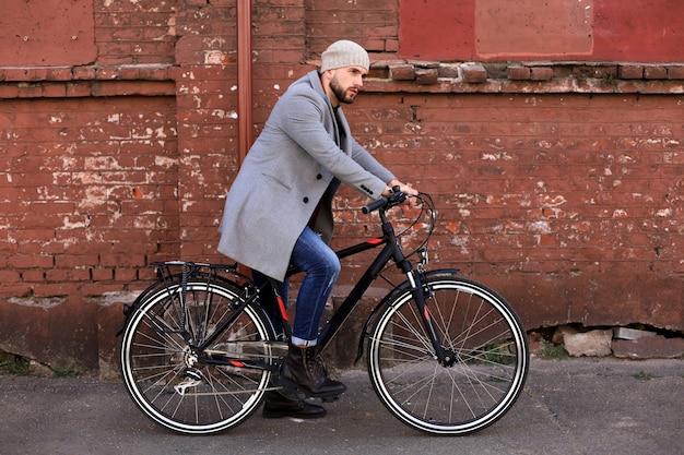 Hübscher junger Mann in grauem Mantel und Hut, der mit seinem Fahrrad steht.