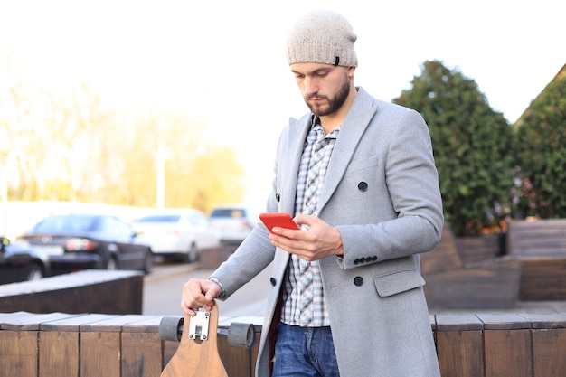 Hübscher junger Mann in grauem Mantel und Hut mit Smartphone, ruhend, stehend mit Longboard. Urbanes Skateboard-Konzept.