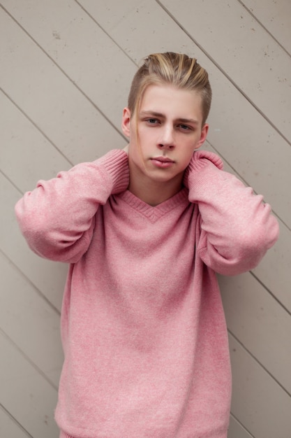 Hübscher junger Mann mit blondem Haar in einem rosa Modepullover nahe einer Holzwand