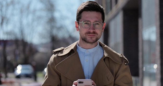 Hübscher junger Mann mit Brille, der in stilvoller Kleidung spazieren geht und ein Smartphone auf der Straße benutzt Mann in der Stadt, der sich mit Menschen verbindet, die online Nachrichten senden und im Internet surfen