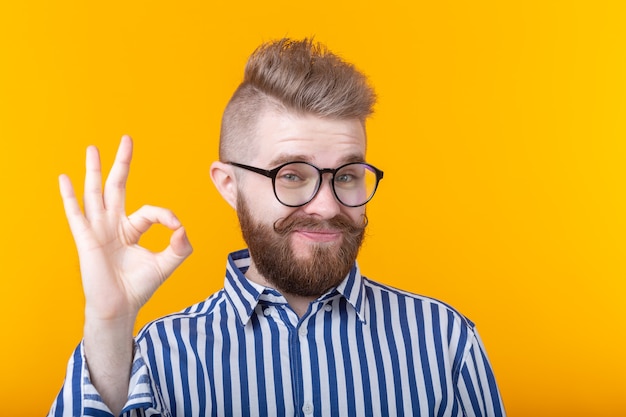 Hübscher junger Mann mit einem Schnurrbart und einem Bart mit Brille zeigt ok posiert auf einer gelben Wand