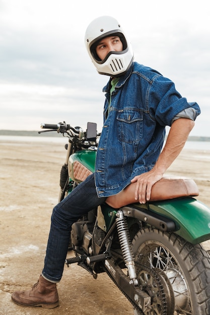 Hübscher junger Mann mit lässigem Outfit, der auf einem Motorrad am Strand sitzt und einen Helm trägt