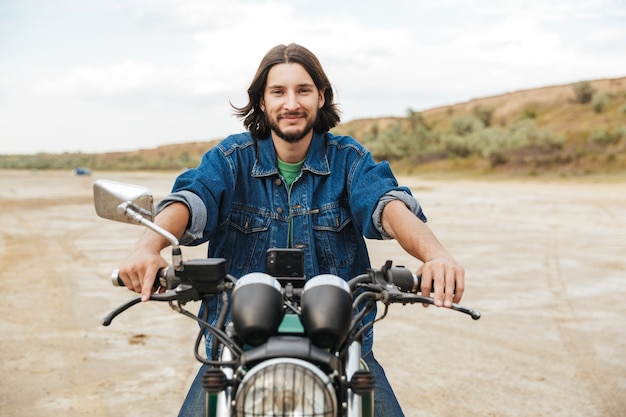 Hübscher junger Mann mit lässigem Outfit, der auf einem Motorrad am Strand sitzt