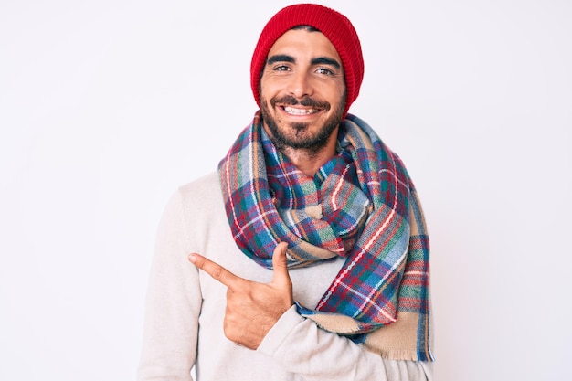 Hübscher junger Mann mit lockigem Haar und Bär, der einen Winterpullover, einen Schal und eine Wollmütze trägt und fröhlich lächelt und mit der Hand und dem Finger zur Seite zeigt