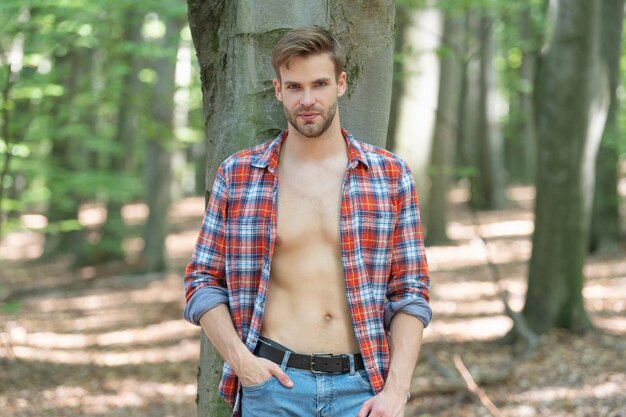 Hübscher junger Mann mit lumbersexuellem Blick in aufgeknöpftem Holzfällerhemd, das am Baum steht