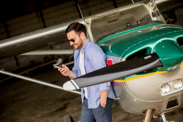 Hübscher junger Pilot, der Flugzeug im Hangar überprüft und Handy verwendet