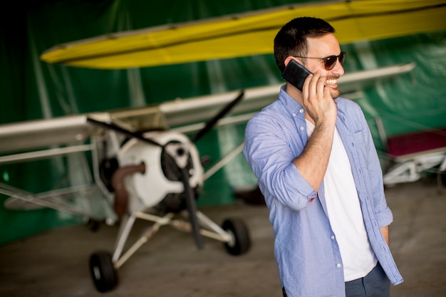 Hübscher junger Pilot, der Flugzeug im Hangar überprüft und Handy verwendet