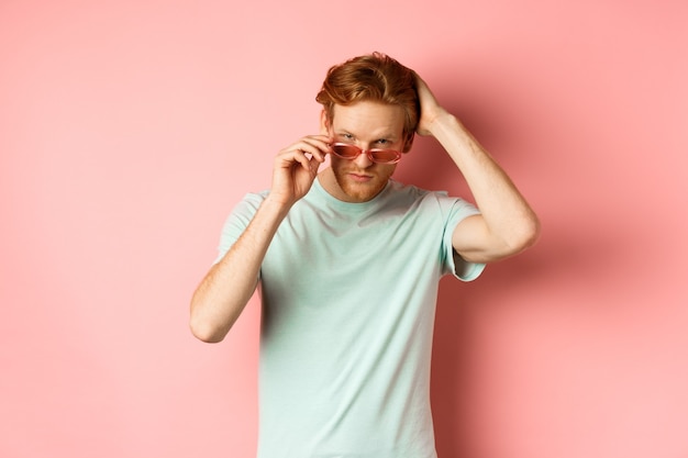 Hübscher junger rothaariger Mann mit Sonnenbrille, der mit der Hand das Haar bürstet und selbstgefällig und selbstbewusst auf c...