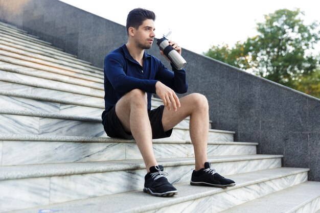 Hübscher junger Sportler, der Wasser von einer Flasche draußen trinkt, auf Treppe sitzt