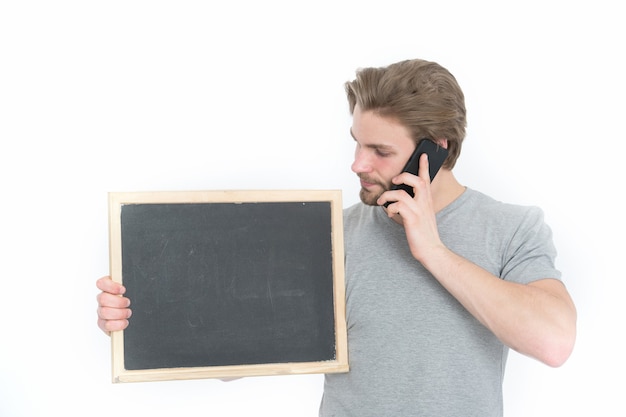 Hübscher junger Student im Hemd benutzt ein Smartphone und lächelt, steht mit Tafel, Kopienraum