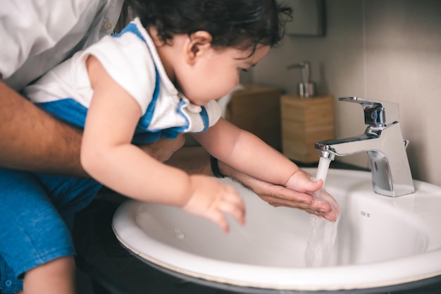 Hübscher junger Vater, der kleine kleine Tochter anhebt, während er sie in Sauberkeit schult, indem er ihr hilft, ihre Hände unter fließendem Leitungswasser im Badezimmerwaschbecken zu waschen