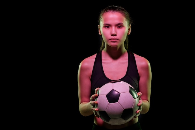 Hübscher junger weiblicher Fußballspieler, der Fußball während des Trainings in Isolation vor der Kamera über schwarzem Hintergrund hält