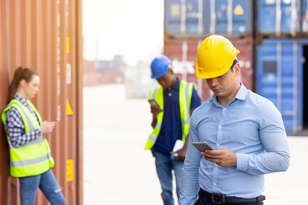 Hübscher kaukasischer Ingenieur und Gruppenarbeiter, der auf digitalem Mobiltelefon in einer Pausenzeit steht, die mit Frachtcontainer im Hintergrund steht