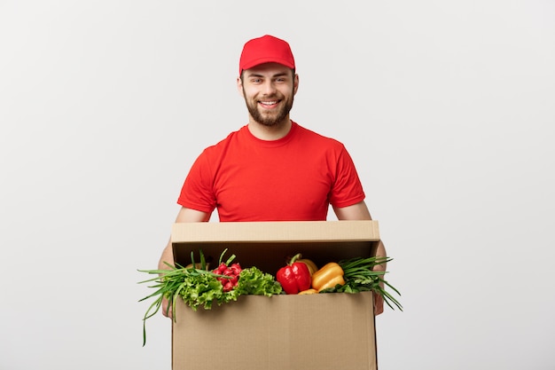 Hübscher kaukasischer Lebensmittellieferungskuriermann in roter Uniform mit Einkaufsbox mit frischem Obst und Gemüse