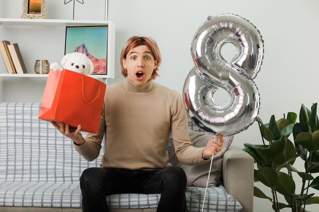 Hübscher Kerl am glücklichen Frauentag, der den Ballon Nummer acht mit der Geschenktüte hält, die auf dem Sofa im Wohnzimmer sitzt