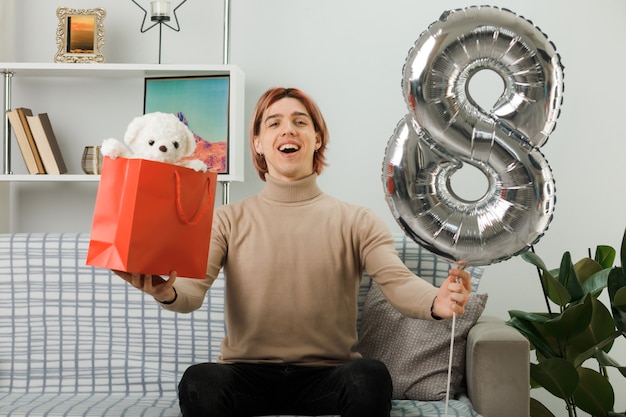 Hübscher Kerl am glücklichen Frauentag, der den Ballon Nummer acht mit der Geschenktüte hält, die auf dem Sofa im Wohnzimmer sitzt