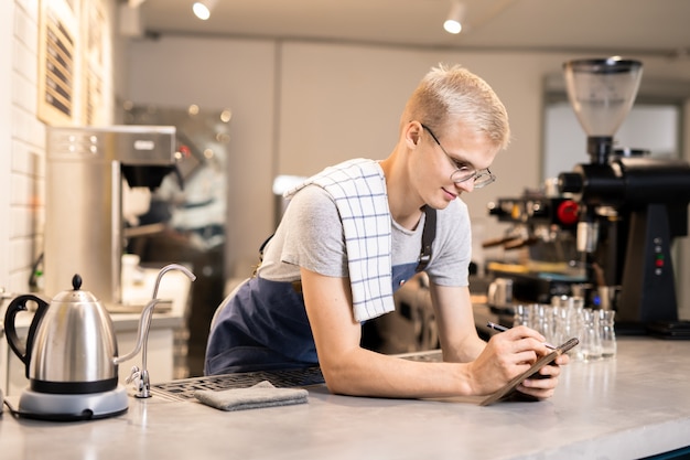 Hübscher Kerl in Schürze und Abschlag, der zu Beginn des Arbeitstages in der Cafeteria Bestellungen in seinem Notizblock durchschaut