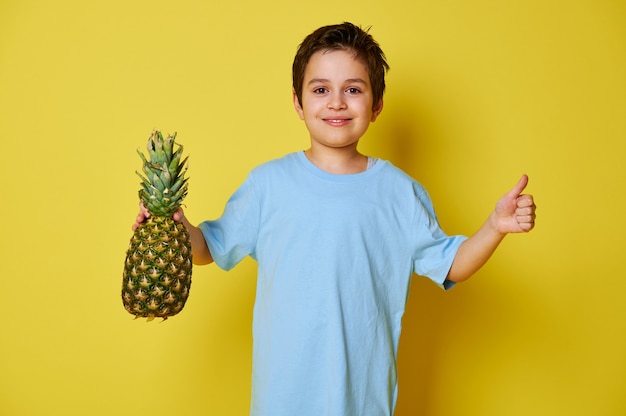 Hübscher Kinderjunge, der eine Ananas hält und Daumen oben zeigt