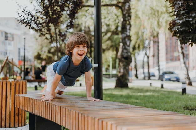 Hübscher kleiner brünetter Junge in einem blauen T-Shirt und weißen Shorts spielt auf einer Holzbank im Park. Kind, das draußen geht. Konzept der Gesundheit. Platz für Text. Foto in hoher Qualität