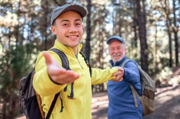 Hübscher kleiner Junge geht glücklich in den Wald und hält die Hand seines Großvaters Lächelnde Mehrgenerationenfamilie, die gemeinsam Freiheit, Berg und Natur genießt Die neue Generation hilft der alten