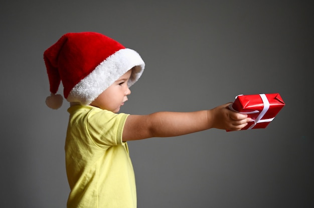 Hübscher Kleinkindjunge, Weihnachtsmütze auf dem Kopf, die Geschenkbox heraushaltend. Weihnachts- oder guten Rutsch ins Neue Jahr-Shopping-Konzept. Foto in hoher Qualität