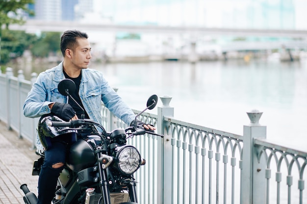 Hübscher kühler asiatischer Mann in der Jeansjacke, die neben Motorrad steht und Fluss betrachtet
